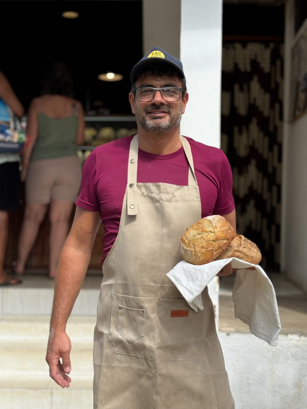 Miquel na porta da padaria, de avental, segurando dois pães.